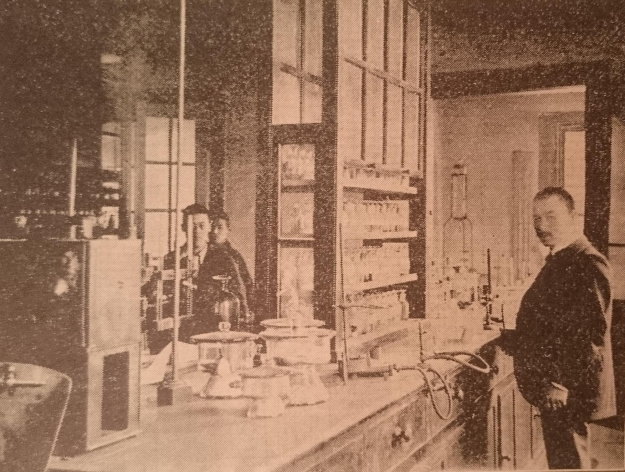 真ん中に試験に使う道具がたくさん並んだ机があり、右側には男性が1人立ち、左奥には男性が2人映しだされた飲食物試験室内の白黒写真