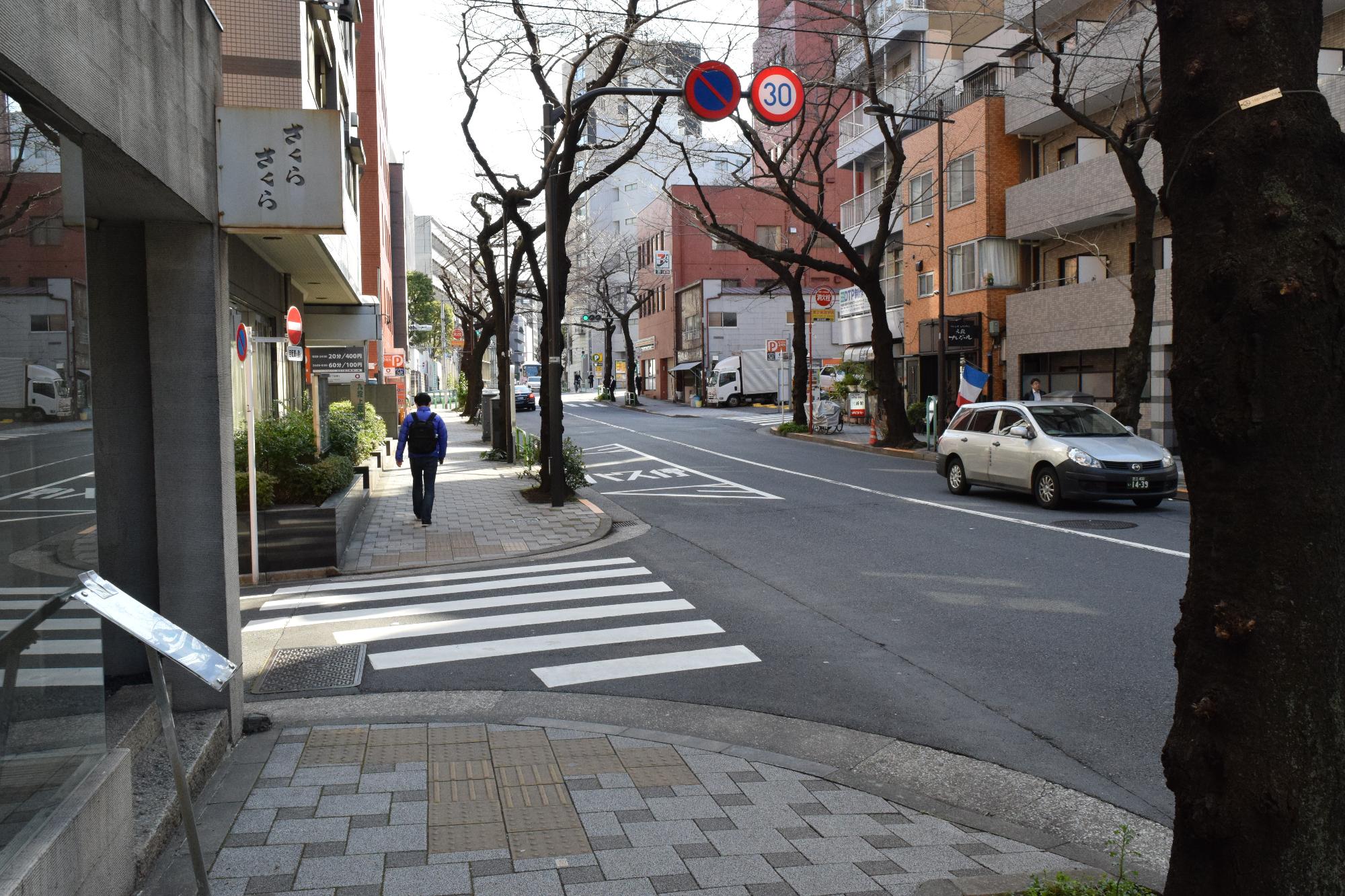 ビルなどの建物が建ち並ぶ住宅街沿いの歩道を歩いている人や、道路を車が走行している様子の写真