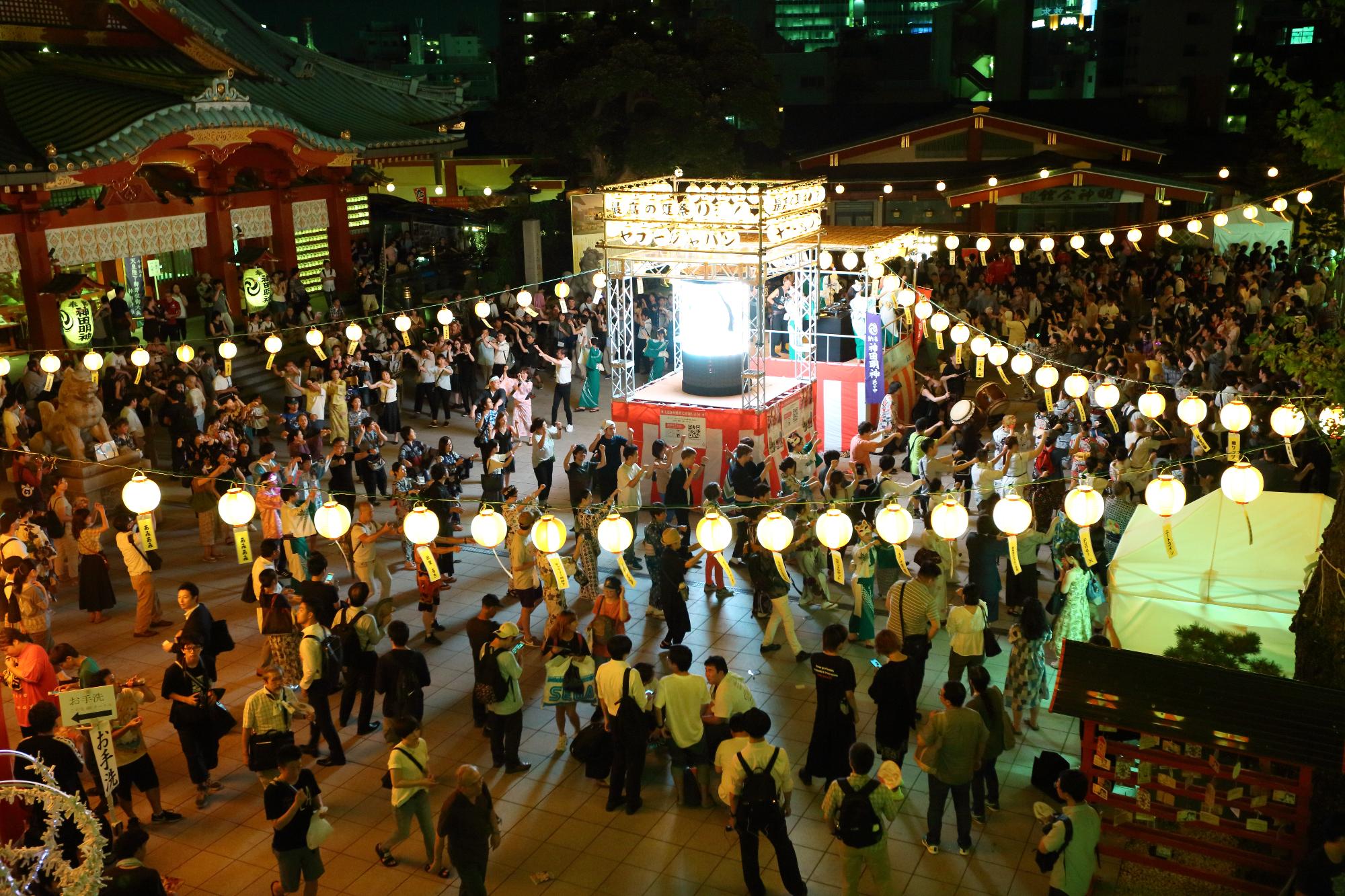 提灯がぶら下がり、紅白幕で覆われたやぐらが設置された祭り会場に沢山の人達が集まっている写真