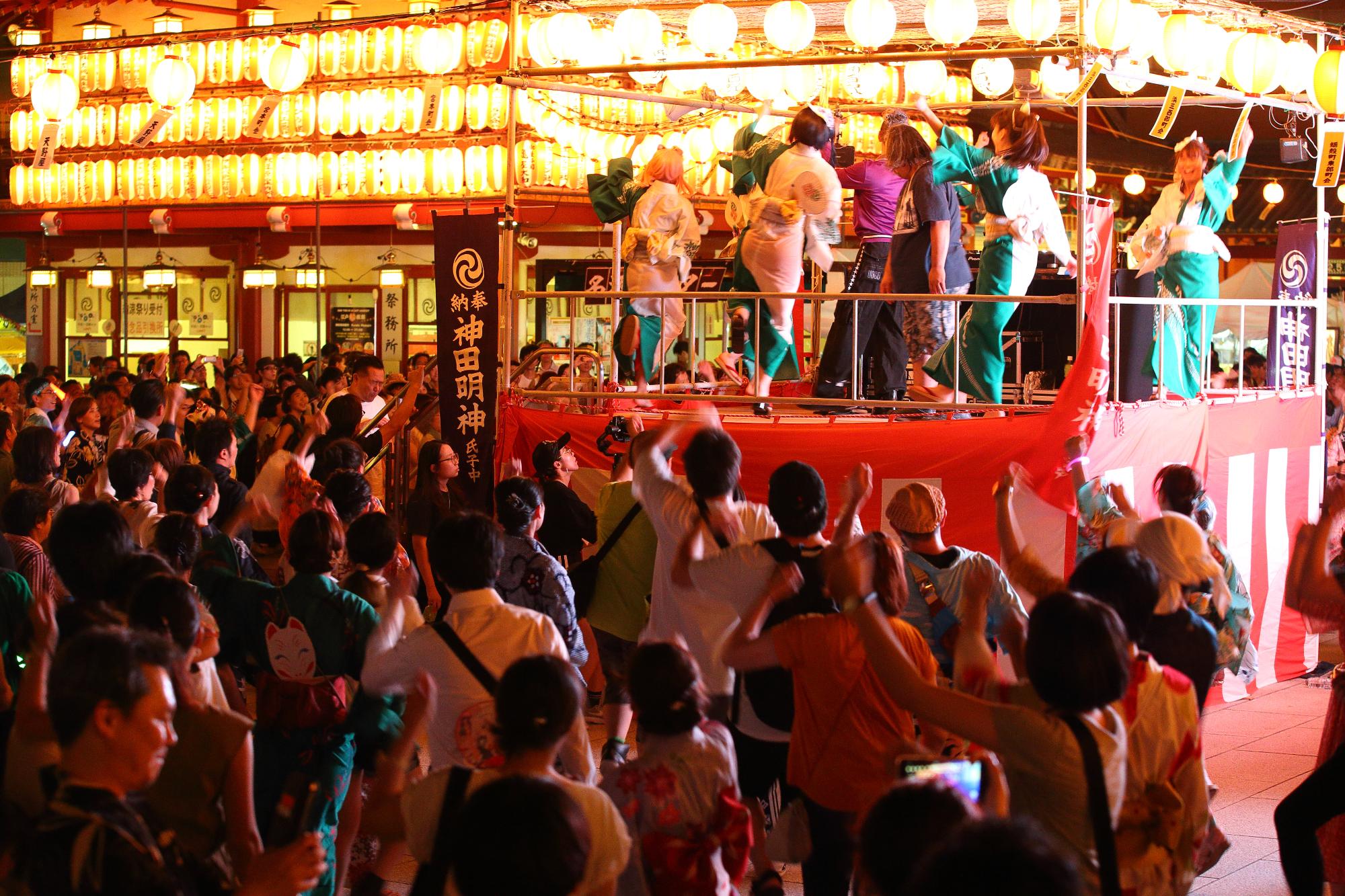 明かりが灯され、紅白幕が巻かれたやぐらの上で踊る方々、その下でやぐらを囲んで踊りを踊っている方々の祭りの様子の写真