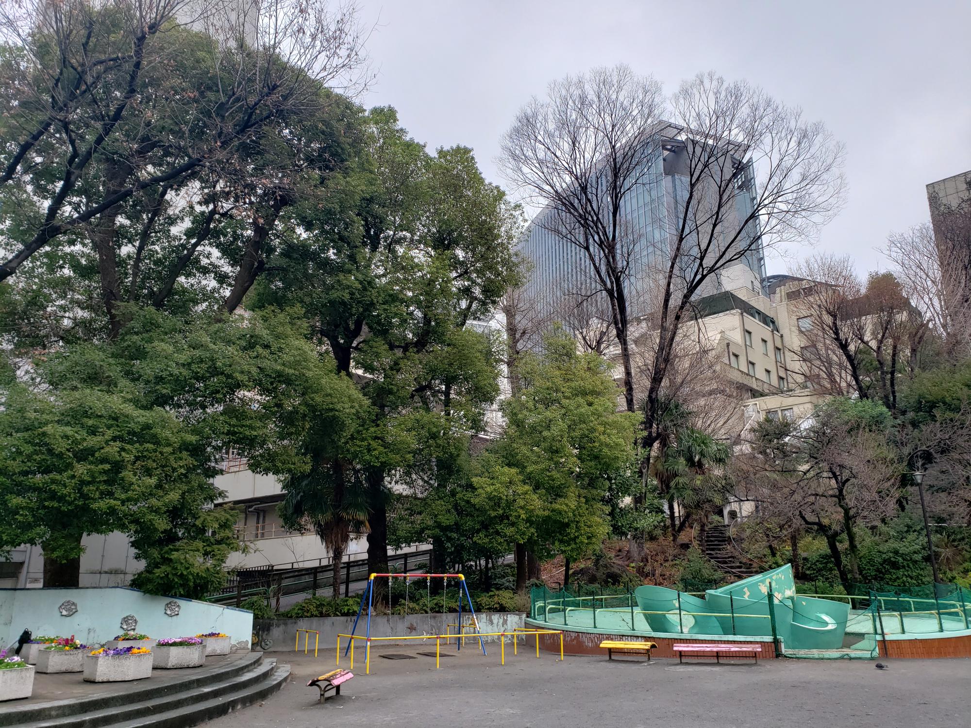 周辺に木々が立ち並び、緑色の滑り台やブランコ、ベンチなどが設置された錦華公園の写真