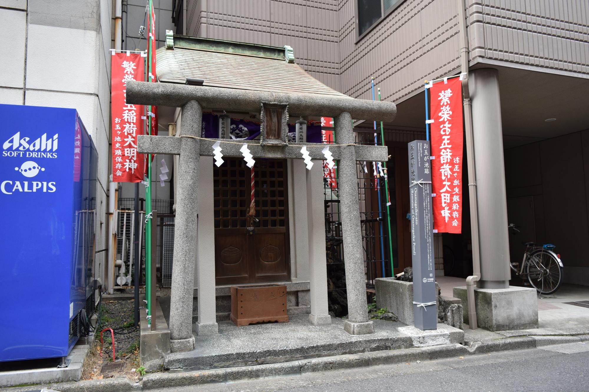 自動販売機と建物の間に建つお玉稲荷神社の写真