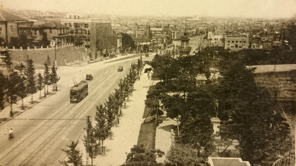 周辺に木々が立ち並び、道路の中央を市電が走っている様子を上空から写した絵葉書の白黒写真